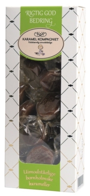 En hvid æske med vindue og på siderne er et sølvnet af små karameller tegnet. I midten flyder en lysegrøn stribe med sirlig tekst: "Rigtig god bedring" - Karamel Kompagniets logo - Uimodståelige bornholmske karameller"